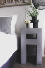 Load image into Gallery viewer, 60cm high square edged concrete bedside table shown in urban grey colour with 10cm shelf. Displayed with foliage on top and a black pendant light above next to a bed dressed with white and grey linen